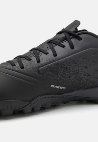 Chaussure de sport noire avec une surface texturée, un design épuré et un logo brillant contrasté. La semelle extérieure présente un motif rugueux pour une meilleure adhérence.