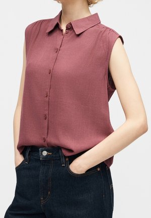 Mouwloze mauve blouse met knopen, ingestopt in donkerblauwe high-waist jeans met handen in de zakken, getoond van nek tot heupen.