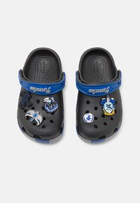 Black Crocs with blue strap, featuring decorative charms: a raven, a crown, and Ravenclaw-themed accents. Ventilation holes throughout.