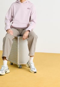 Felpa con cappuccio rosa chiaro, dotata di tasca frontale, abbinata a pantaloni a righe beige. Il modello indossa sneakers grigie e calzini bianchi, seduto su un oggetto metallico.