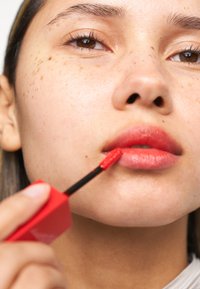 Röd läppglans som appliceras med en applikator, med en fyrkantig röd applikator, glansig finish på läpparna, subtila fräknar och neutral hudton.