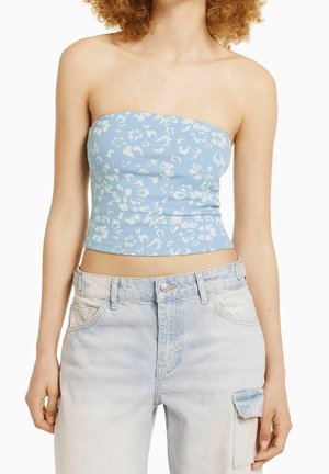 Frau trägt ein hellblaues, florales trägerloses Crop-Top und hoch taillierte helle Jeans mit einer Cargotasche am rechten Bein.
