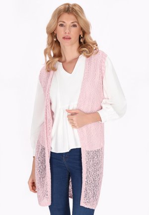 Femme portant un chemisier blanc et un gilet tricoté rose sans manches ouvert devant, associé à un jean bleu foncé, debout devant un fond uni.