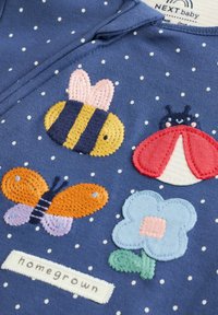 Marinblått tyg med vita prickar pryds av färgglada broderade motiv: en bi, nyckelpiga, fjäril, blomma och en etikett med texten "hemodlad".