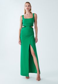 Sieviete, tērpusies zaļā, bez piedurknēm kleitā ar izciļņiem vidukļa rajonā un augstu šķēlumu, stāvot pret vienkāršu gaišu fonu.