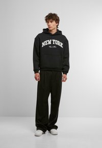 Zwarte hoodie met "NEW YORK" opdruk, relaxte pasvorm, voorzak, gecombineerd met losse zwarte broeken en witte sneakers.