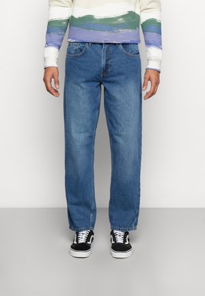 Mann trägt blaue Jeans mit geradem Bein, schwarz-weiße Turnschuhe und einen weißen Pullover mit grünen und lila horizontalen Streifen.