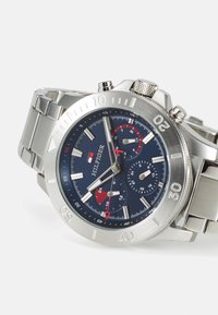 Silberner Edelstahl-Chronograph mit einem dunkelblauen Zifferblatt, roten Akzenten, drei kleinen Zifferblättern und einer strukturierten Krone. Markierte Lünette mit Minutenanzeigen.
