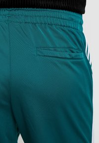 Türkisfarbene Sporthosen mit glatter Textur, elastischem Bund und einer Gesäßtasche. Besitzt weiße Streifen an den Seiten.