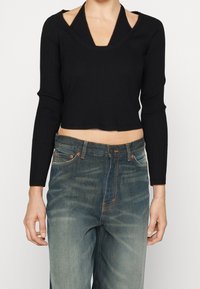 Schwarzes, geripptes, kurzes Top mit V-Ausschnitt und offenen Schulterausschnitten, kombiniert mit ausgebeulten blauen Jeans mit orangefarbenen Nähten und vorderen Taschen.