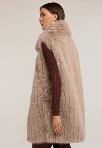 Beżowa kamizelka z futra sztucznego z wysokim kołnierzem, charakteryzująca się miękką, puszystą teksturą i luźnym krojem, noszona na brązowej bluzce z długim rękawem.