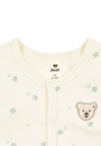 Kremowy dziecięcy kombinezon z jasnooliwkowymi wzorami koniczyny, brązową haftowaną twarzą misia na piersi oraz zatrzaskami z przodu. Rozmiar 2-3 miesiące.