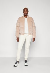 Giacca puffer rosa chiaro con colletto alto e logo a patch, abbinata a pantaloni bianchi con dettagli laterali e sneakers grigie.