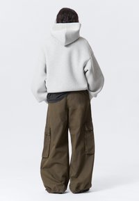 Persona che indossa un ampio hoodie grigio chiaro e pantaloni cargo larghi di un verde oliva scuro, in piedi con le mani in tasca, girato di spalle su uno sfondo semplice.
