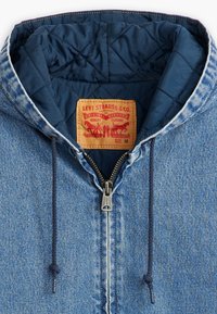 Veste en denim à capuche avec une doublure matelassée navy. Comprend une fermeture éclair en métal, des cordons de serrage et une étiquette marron avec des graphiques rouges. Taille M.