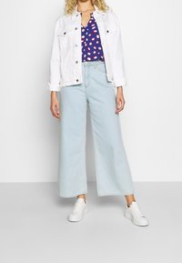 Vit denimjacka, blå topp med röda prickar, ljusblå vida jeans, vita sneakers. Fokus på avslappnad, lager-på-lager outfitdesign.