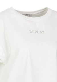 Camiseta blanca de algodón con cuello redondo y mangas cortas, con la palabra "REPLAY" impresa en plata en el frente. Textura suave.