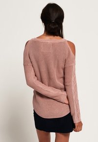 Rosa Strickpullover mit offenen Schultern, strukturierendem Muster an den Ärmeln, geripptem Design und abgerundetem Saum. Getragen über einem schwarzen Rock.