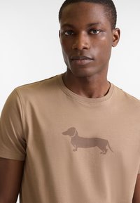 Camiseta color beige de manga corta con un discreto gráfico de un dachshund marrón oscuro centrado en el pecho, hecha de una tela suave y liviana.