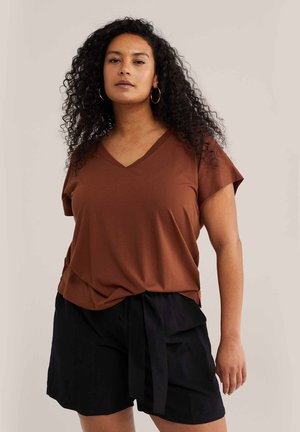 Femme aux cheveux bouclés portant un haut marron à col en V et un short noir avec une ceinture nouée, debout devant un fond clair uni.