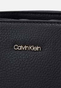Juodas tekstūruotas odinis krepšys su auksiniu „Calvin Klein“ logotipu. Dizainas pasižymi subtiliais grūdų raštais ir lygiu paviršiumi.