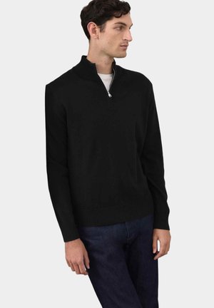 Uomo che indossa un maglione nero con zip al collo sopra una camicia bianca e jeans scuri, in piedi contro uno sfondo chiaro e semplice, che guarda di lato.
