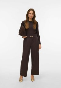 Haut marron à manches longues avec un col texturé, associé à un pantalon marron taille haute à jambes larges et des talons à imprimé serpent, tenant un sac.