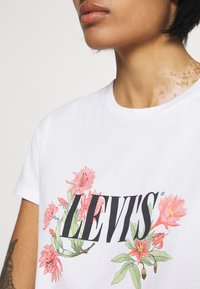 Detailní záběr na osobu v bílém tričku Levi's s růžovými květinovými vzory kolem názvu značky, která ukazuje kožní onemocnění na krku.