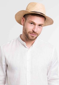Sombrero de paja con ala ancha, color natural y banda oscura. El modelo lleva una camisa de lino blanca con cuello mao. Textura suave.