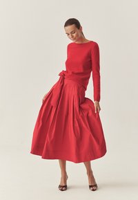 Roter Pullover mit langen Ärmeln, kombiniert mit einem roten, plissierten Midirock mit gebundenem Bund. Der Rock hat eine glatte Textur und einen sanften Fall.