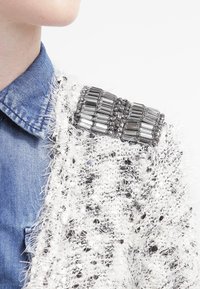 Vit och svart texturerad cardigan med en fluffig stickning. Har en dekorativ brosch med silver- och svarta metallaccent på axeln.