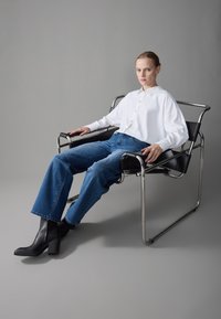 Black leather chair with chrome frame. Model in white shirt, blue jeans, and black heeled boots, seated with hands on chair arms.