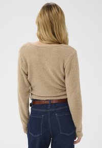 Bež majica s teksturo, dolgimi rokavi in ohlapnim krojem, širokim izrezom, v kombinaciji z temno modrimi kavbojkami, ki imajo dva zadnja žepa in rjav pas.