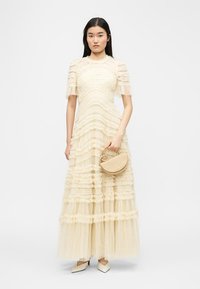 Γυναίκα με φόρεμα από κρέμ τούλι σε στρώσεις, κρατώντας μια μπεζ τσάντα σε σχήμα ημισελήνου, φορώντας μυτερά τακούνια, στέκεται μπροστά σε λευκό φόντο.