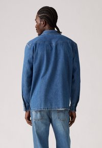 Veste en denim bleu moyen avec des manches longues, poignets à boutons et une coupe droite. Présente un empiècement au dos et des détails de couture subtils.