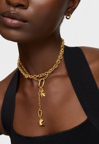 Złoty, wielowarstwowy naszyjnik z wydłużonymi ogniwami, wyrazistym wisiorkiem i wypolerowanymi akcentami, noszony na nagiej skórze na tle czarnej bluzki.