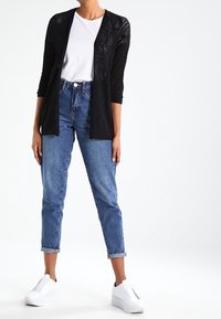 Person wearing black open cardigan, white t-shirt, rolled-up blue jeans, and white sneakers, standing on a white background.