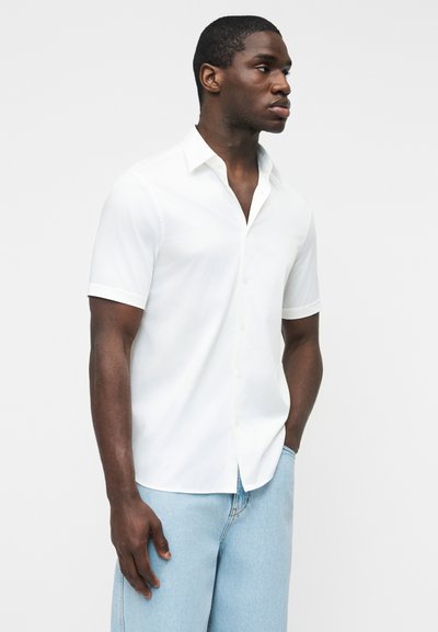 Άνδρας που στέκεται με τα χέρια στις τσέπες, φορώντας λευκό πουκάμισο με κοντά μανίκια και ανοιχτόχρωμο μπλε τζιν, μπροστά σε απλό φόντο.