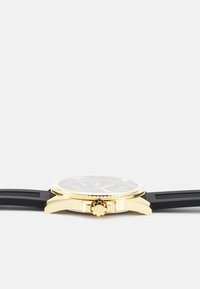 Montre dorée avec un cadran rond, bracelet en caoutchouc noir, lunette texturée et couronne. Verre transparent et cadran noir avec marquages blancs.