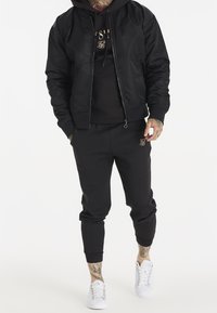 Giacca bomber nera sopra un hoodie nero, abbinato a pantaloni jogger neri. Presenta dettagli dorati sull'hoodie e sui jogger. Sneakers bianche completano il look.