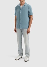 Lichtblauw gemêleerd overhemd met korte mouwen en knoopsluiting, gecombineerd met lichtblauwe jeans en witte sneakers met grijze accenten.