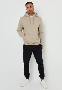 Sudadera beige con cordones, bolsillo canguro y puños de canalé, combinada con pantalones cargo negros que cuentan con múltiples bolsillos y puños elásticos.