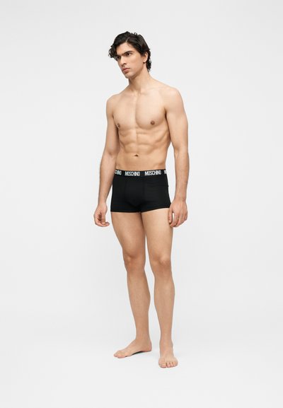 Μυώδης άνδρας στέκεται ξυπόλυτος φορώντας μαύρα μποξεράκια Moschino με λωρίδα μέσης με το λογότυπο, πάνω σε απλό ανοιχτό φόντο.