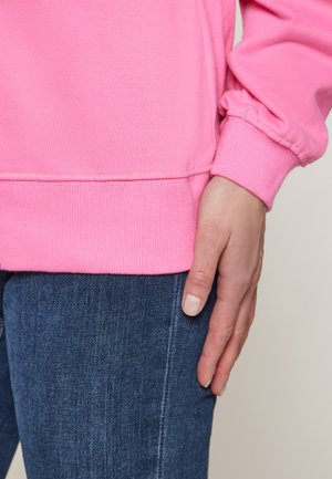 Felpa rosa brillante con polsini e orlo a coste, abbinata a jeans blu scuro. Mano appoggiata sulla coscia, che mostra le trame del tessuto.