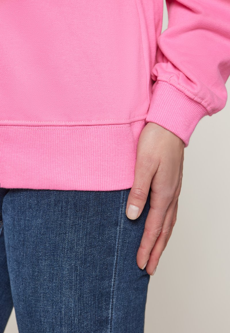 Felpa rosa brillante con polsini e orlo a coste, abbinata a jeans blu scuro. Mano appoggiata sulla coscia, che mostra le trame del tessuto.