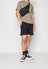 Shorts de sport noirs, t-shirt beige et baskets noires avec semelles blanches. L'individu porte un sac bandoulière noir. Design minimaliste.