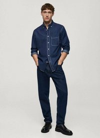 Temno modra denimska srajca z gumbi in žepom na prsih, dolgi rokavi in kontrastno šivanje, v kombinaciji s temnon denim hlačami in črnimi čevlji.