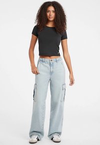 T-shirt nera corta abbinata a jeans cargo a gamba larga azzurri con tasche laterali e chiusura con bottoni, indossata con sneakers bianche.