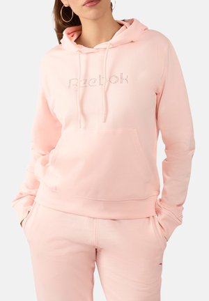 Vrouw draagt een lichtroze Reebok-hoodie en bijpassende joggingbroek met handen in de zakken, getoond van midden bovenbeen tot kin.