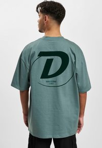 Lys grønn oversized T-skjorte laget av mykt stoff, med en stor mørkegrønn "D" logo og tekst på baksiden. Korte ermer, avslappet passform.